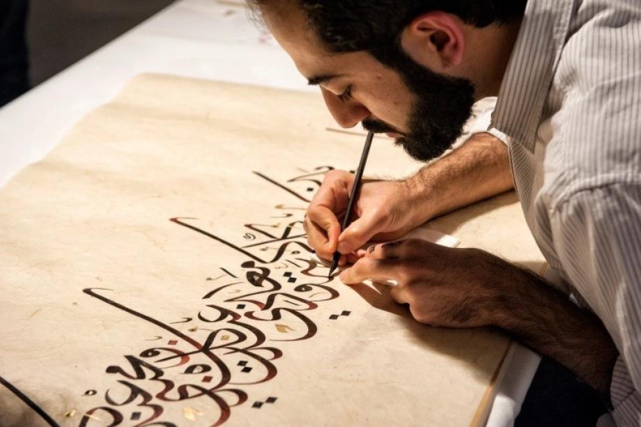 A calligrapher's hand drawing flowing Arabic script with a reed pen