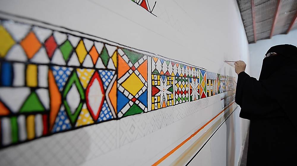 A Saudi artist painting Al-Qatt Al-Asiri geometric patterns across a curved white wall
